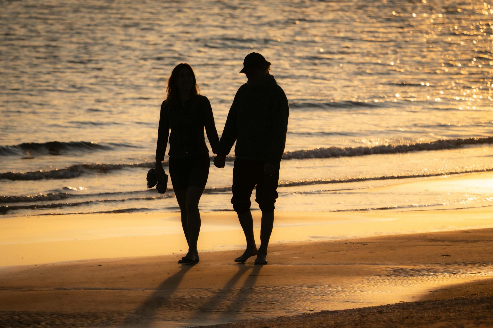 A couple holding hands walking along the shore, silhouetted in golden light