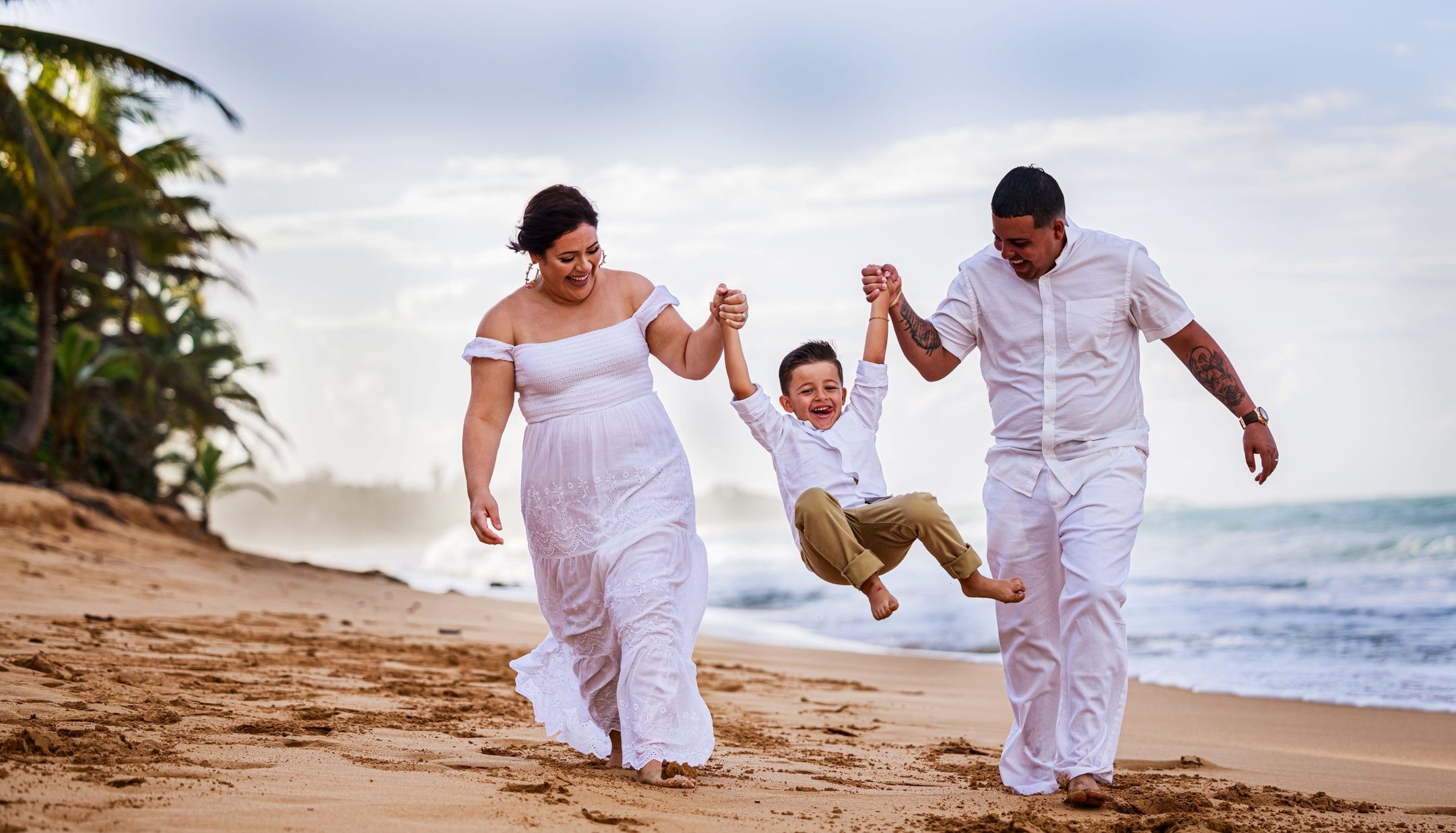 Parents dressed in white swinging their young son by the hands along a sandy shore