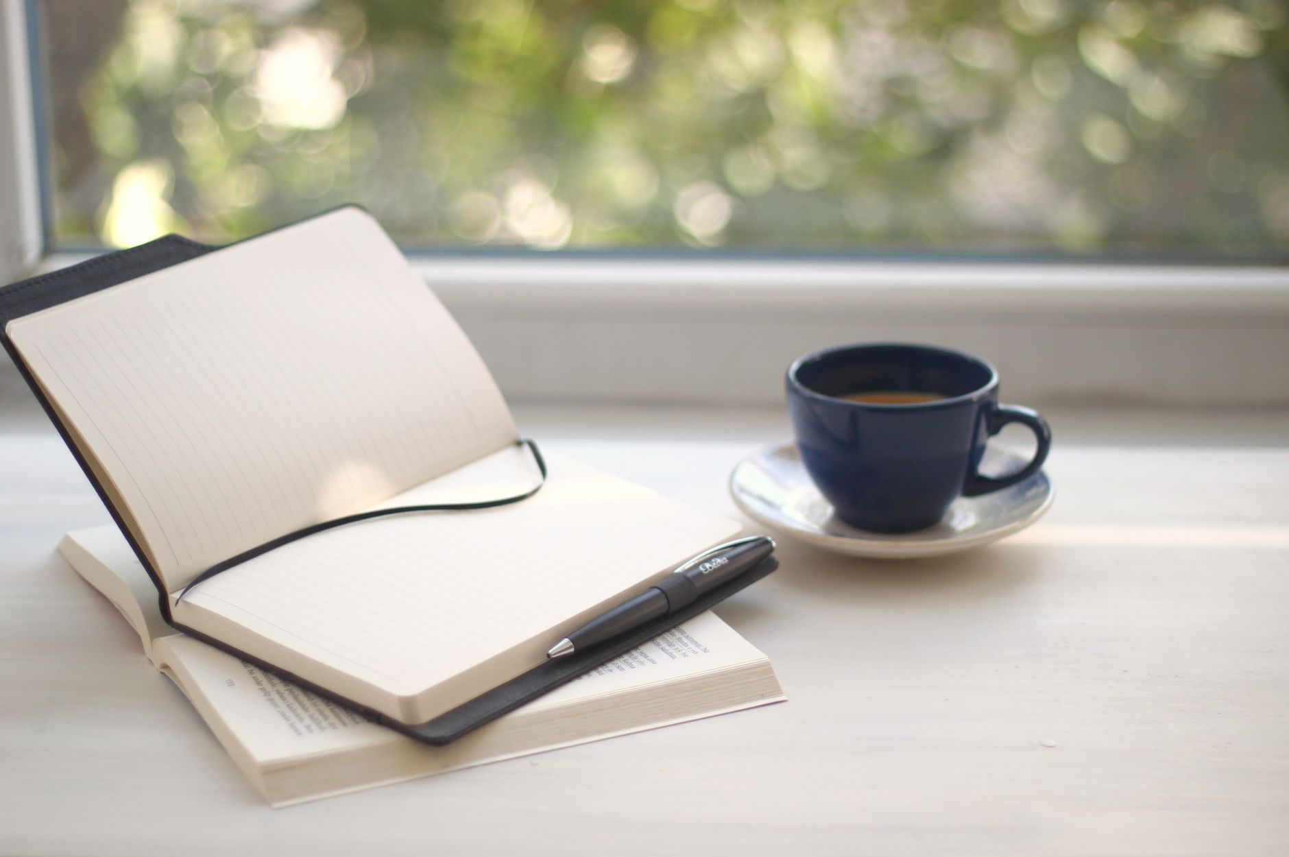 An open blank journal, a pen and a cup of coffee on a sunlit windowsill