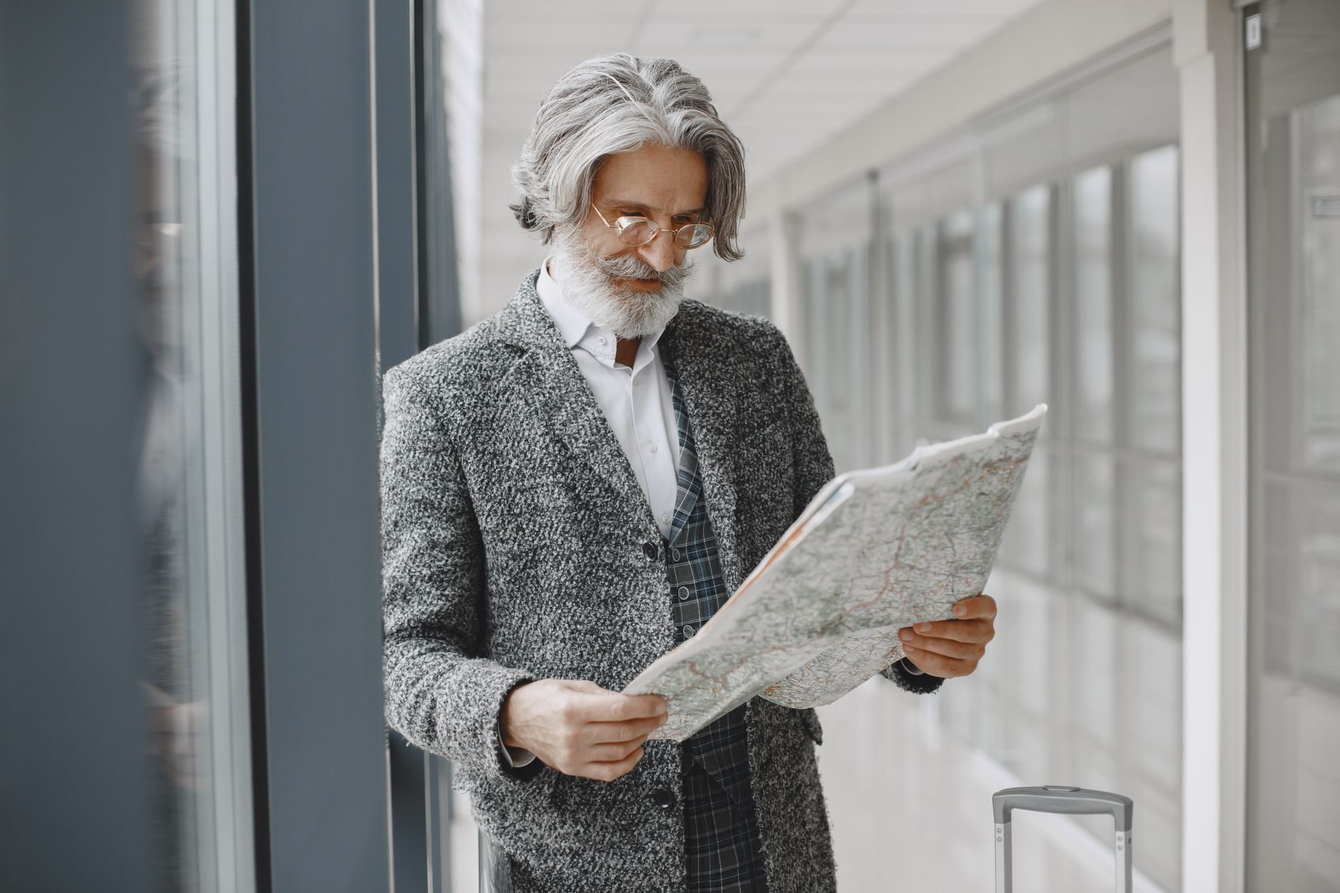 An older bearded man in a gray blazer reads a paper map indoors near large windows