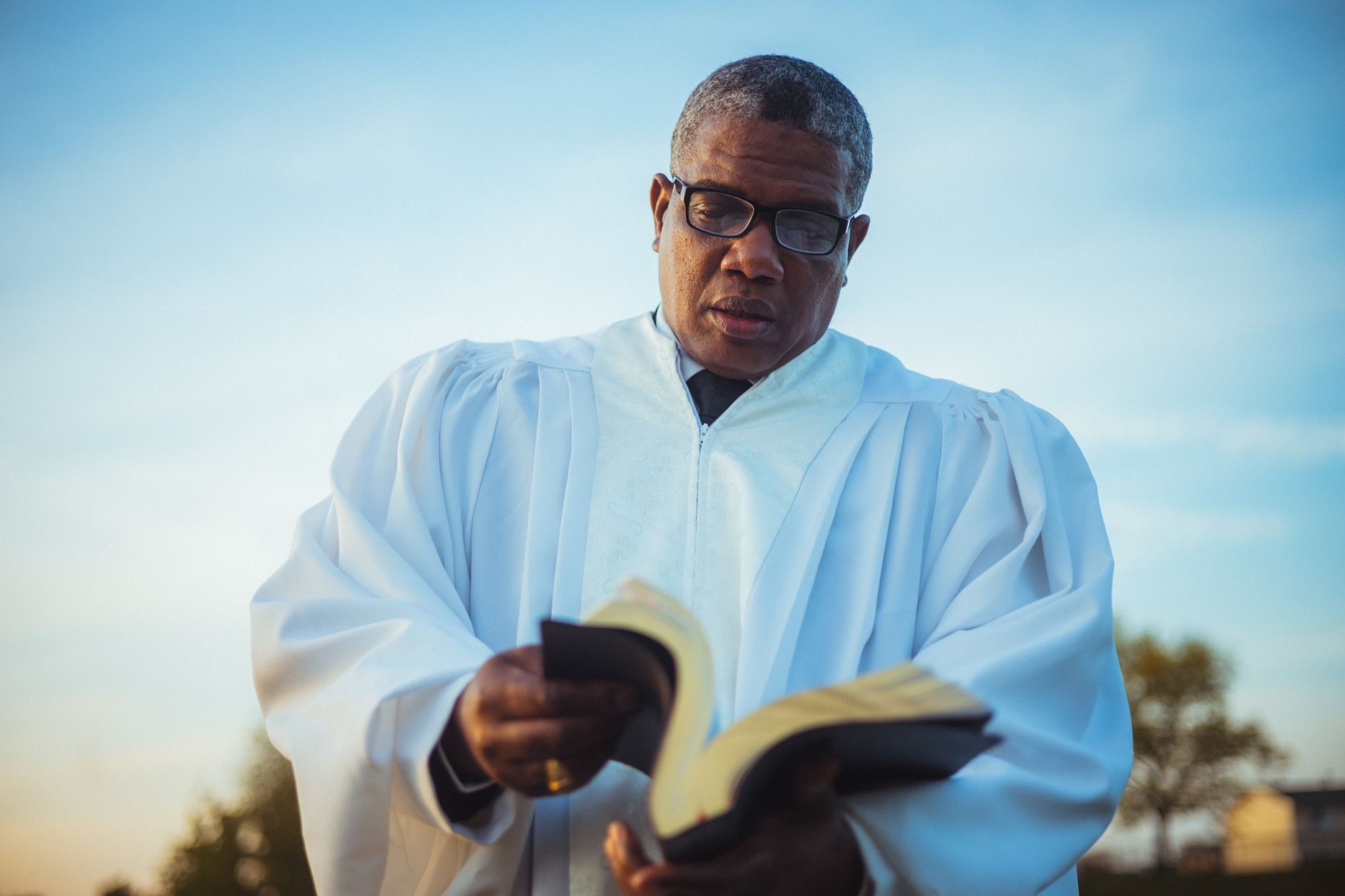 A man in a white robe reads an open book outdoors under a soft blue sky