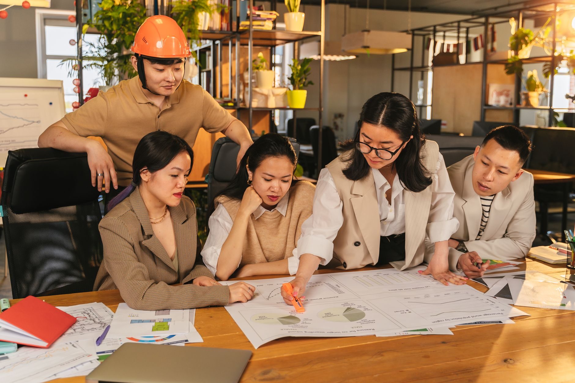 Five people gathered around a wooden table reviewing large printed documents indoors