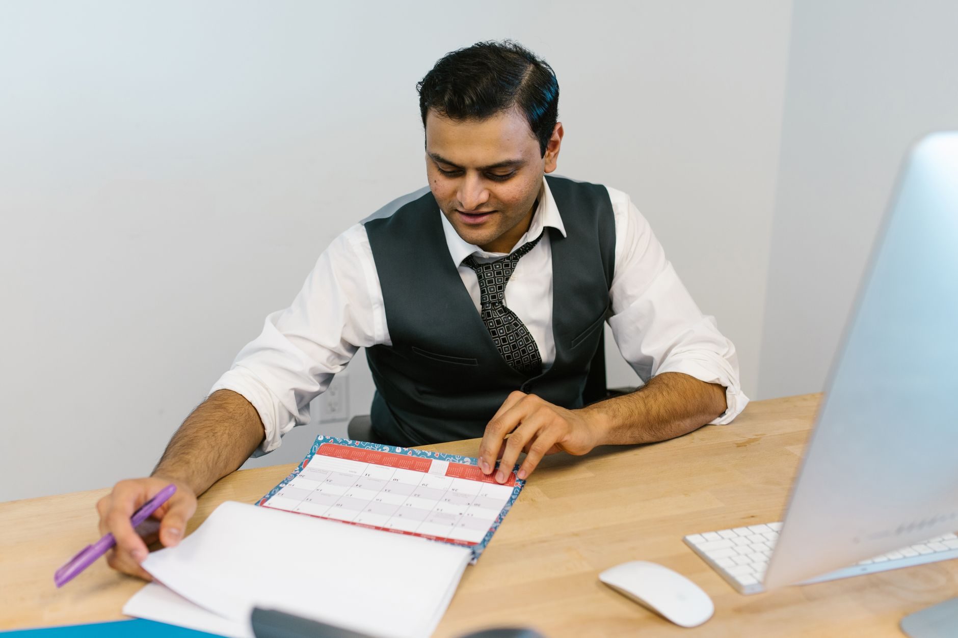 A man in a vest and tie writes notes beside a desk calendar under soft indoor light