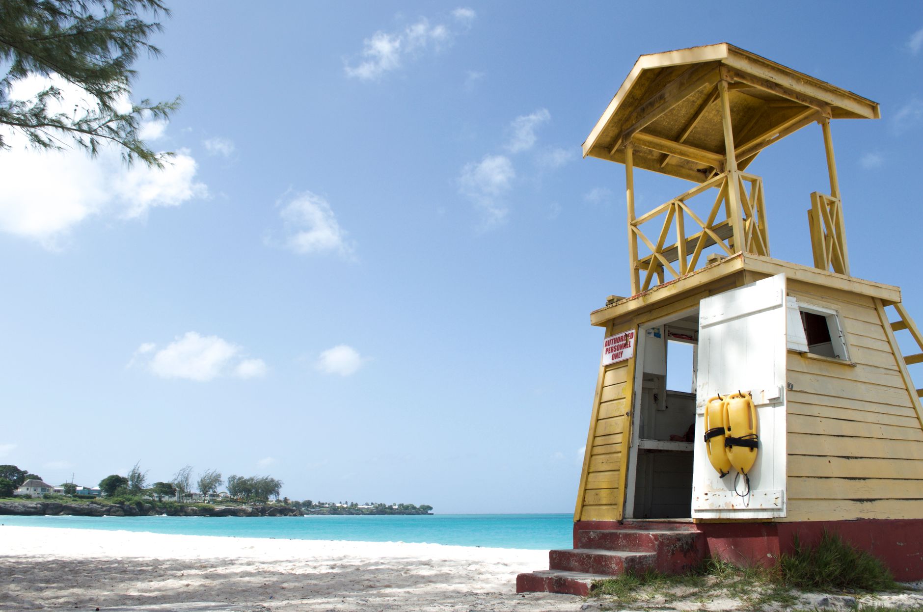 A yellow wooden lifeguard tower with a flotation device stands on a sunny white-sand beach