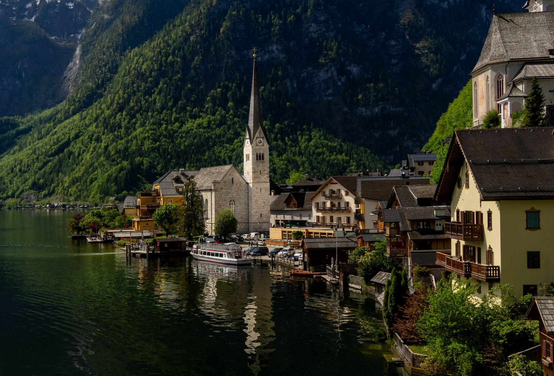 A stone church steeple rises above lakeside buildings beneath forested mountains in dappled sunlight