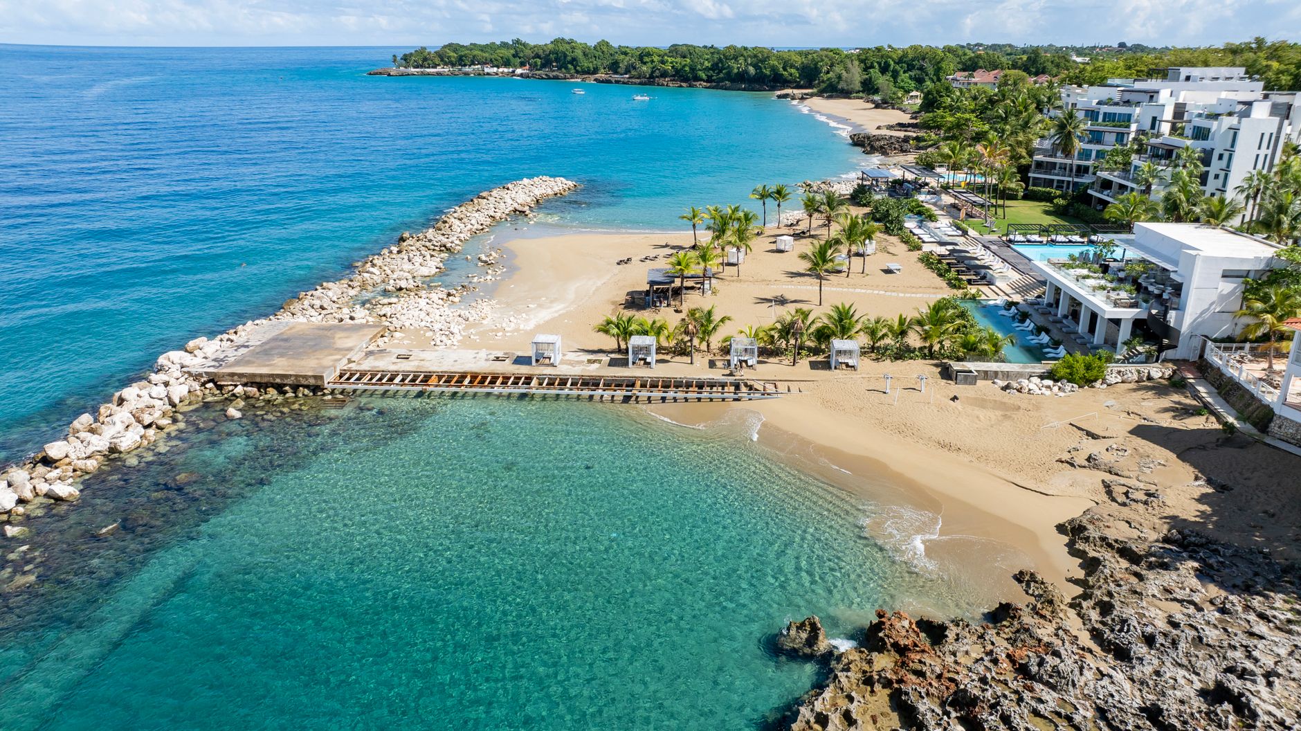 Aerial view of a sandy beach, pier, turquoise water, palm trees, and white resort buildings
