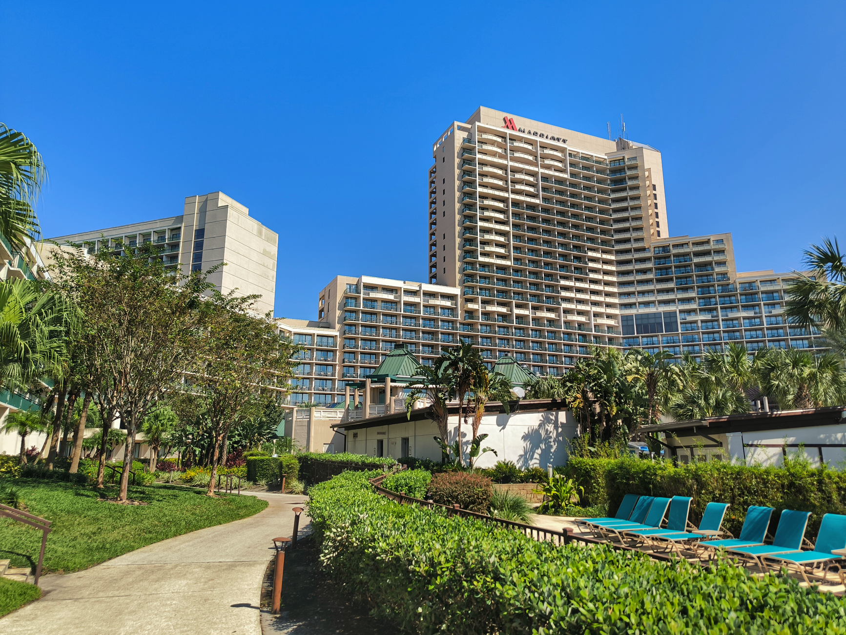 A Marriott hotel tower rises above tropical landscaping, lounge chairs, and a winding pathway in bright sunlight