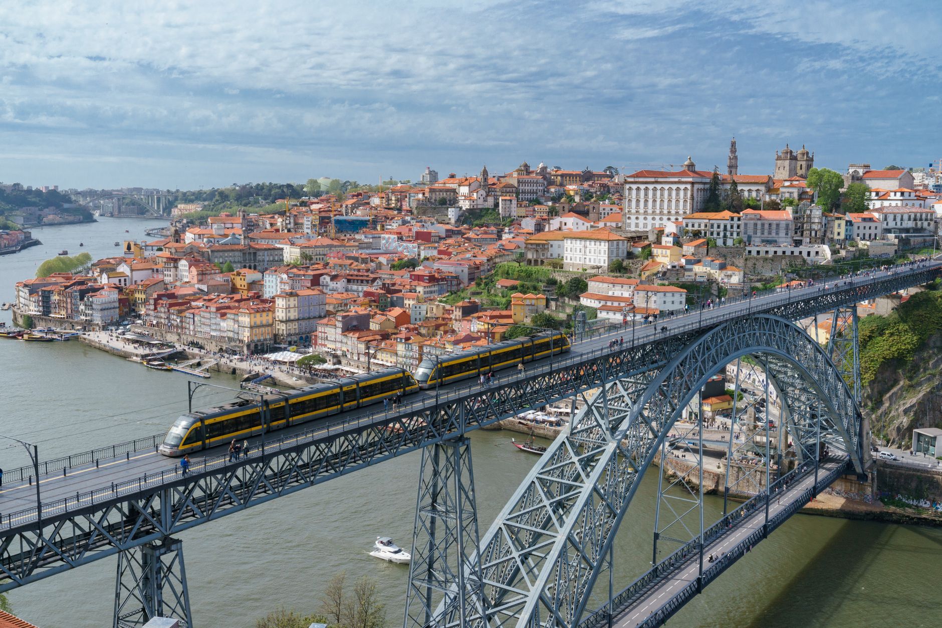 A yellow metro train crosses a large iron arch bridge over a wide river beside a historic hillside city under partly cloudy skies