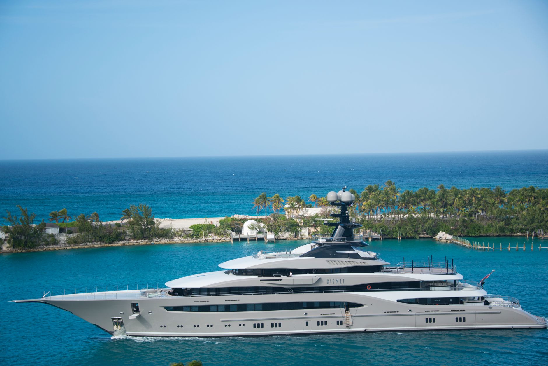 A large white superyacht moves through turquoise water near a palm-lined island under bright sunlight