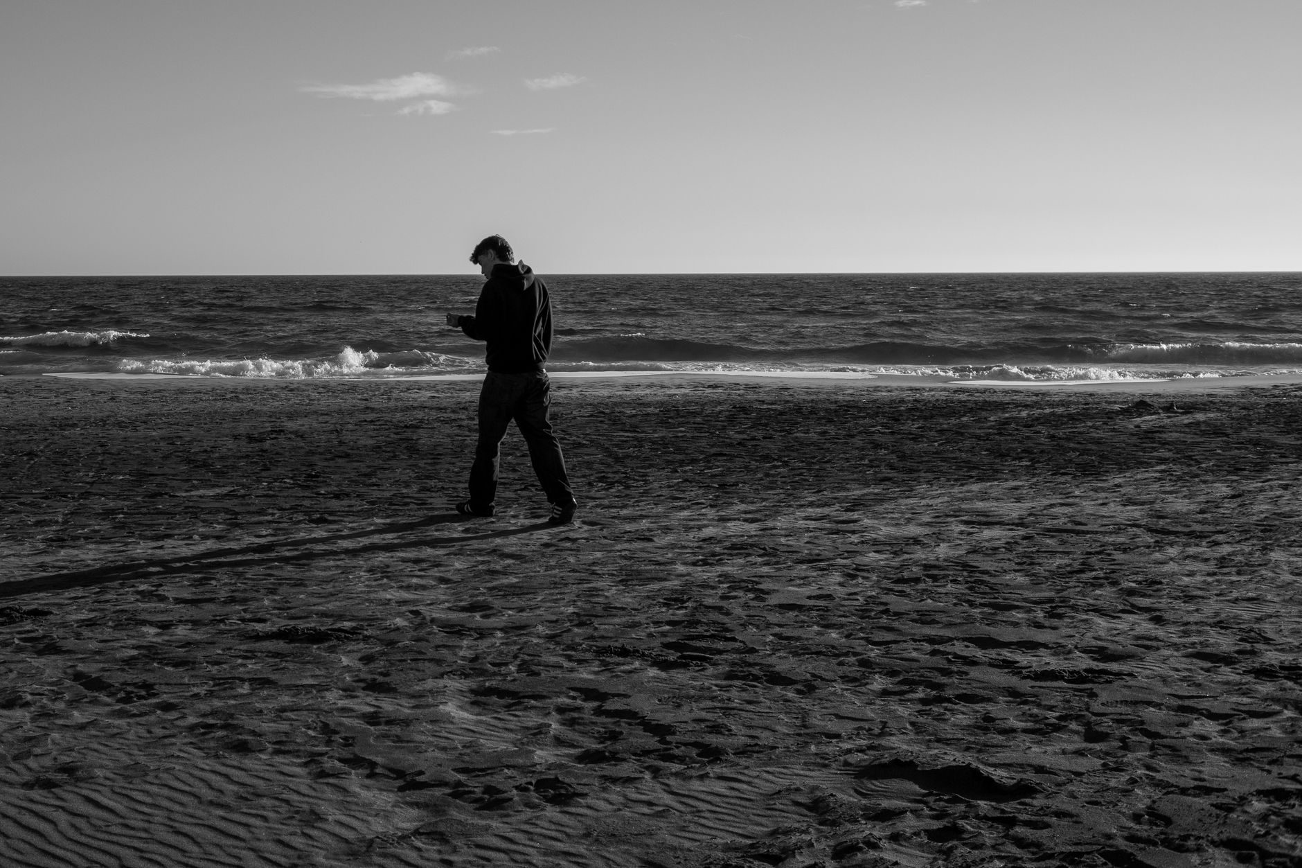 A person walks along a sandy beach holding an object, ocean waves behind them