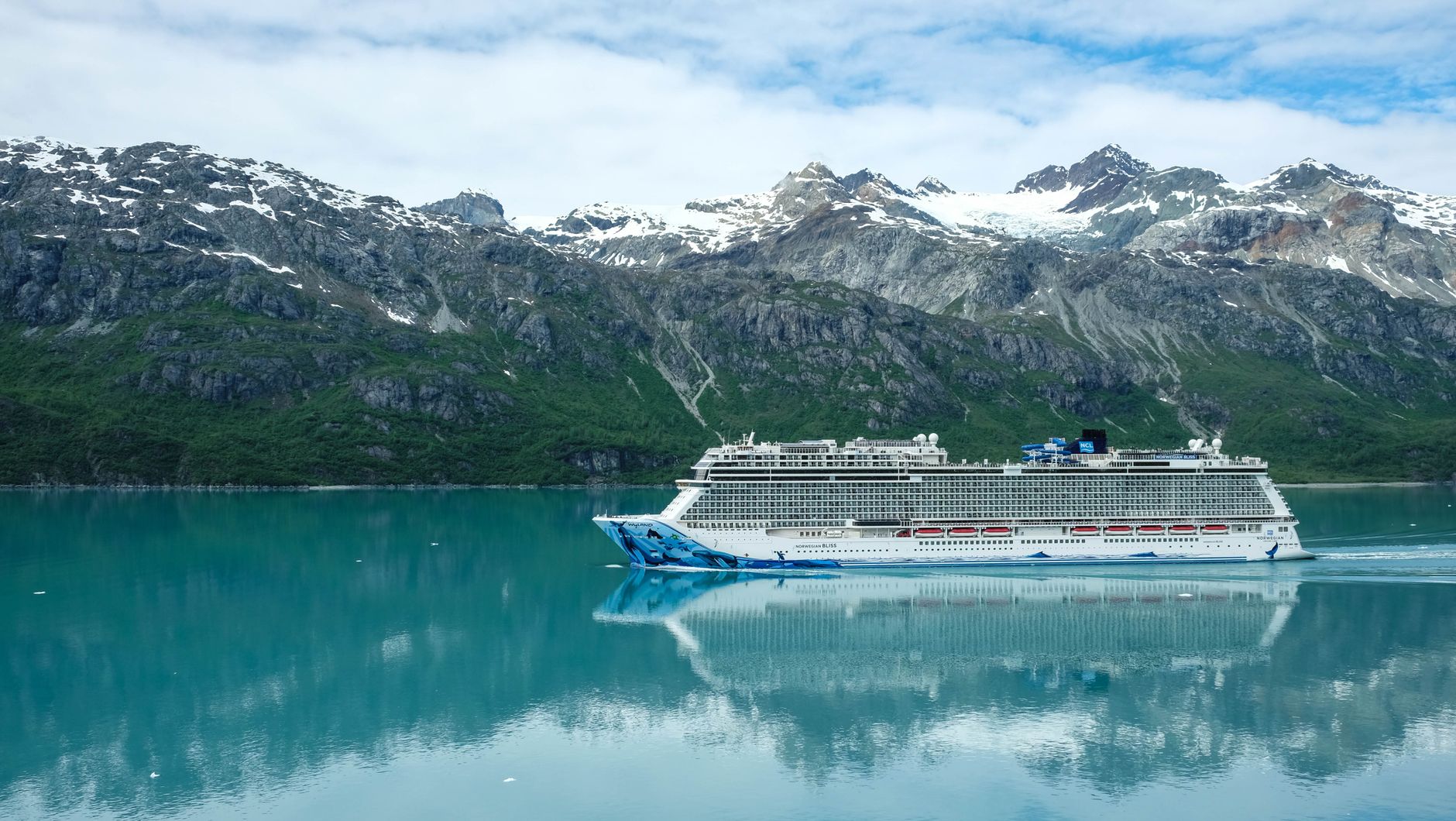 A cruise ship in an Alaskan fjord below forested mountains and ice