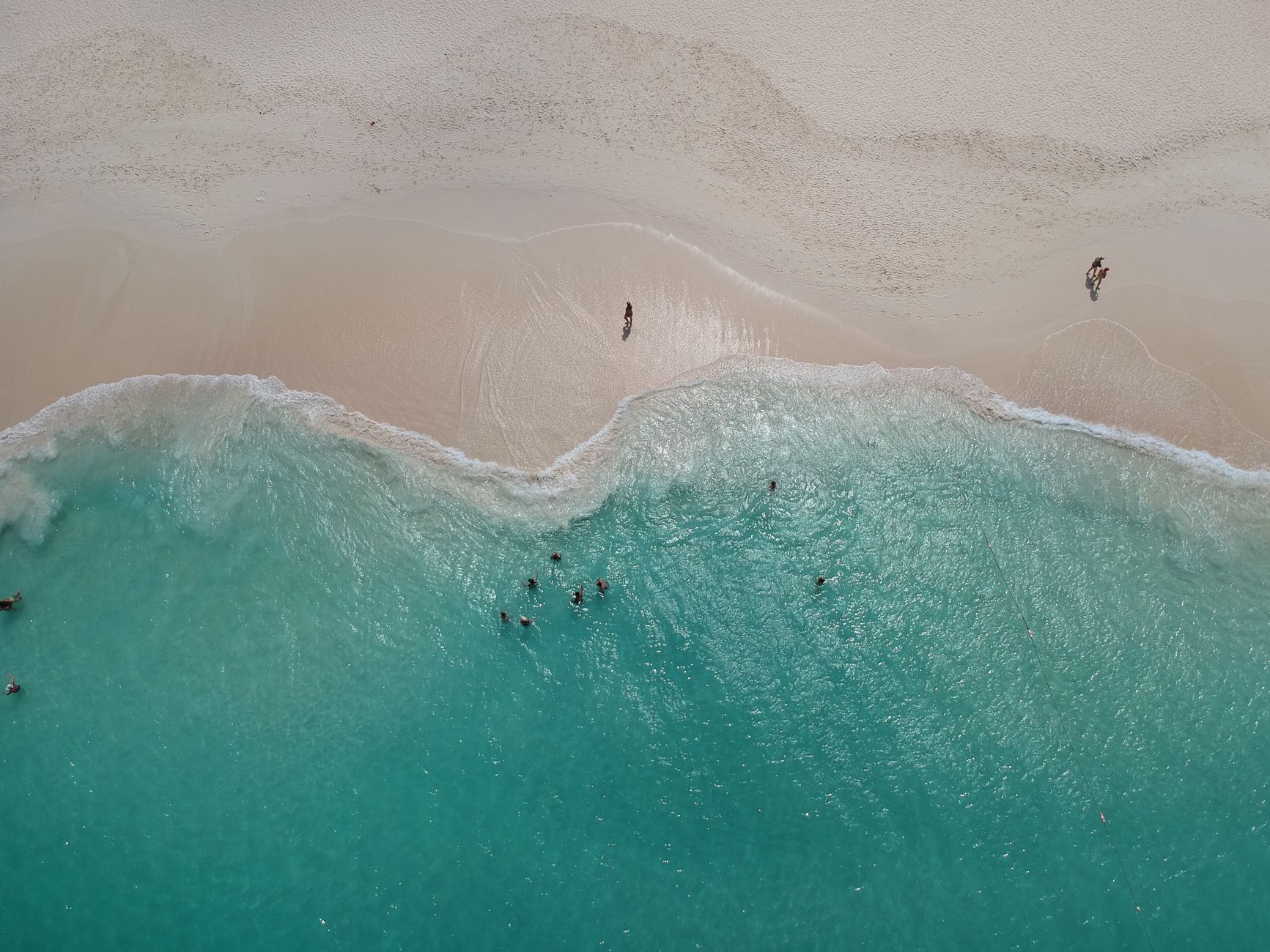 Aerial view of surf and clear green shallows meeting a broad pale sand beach