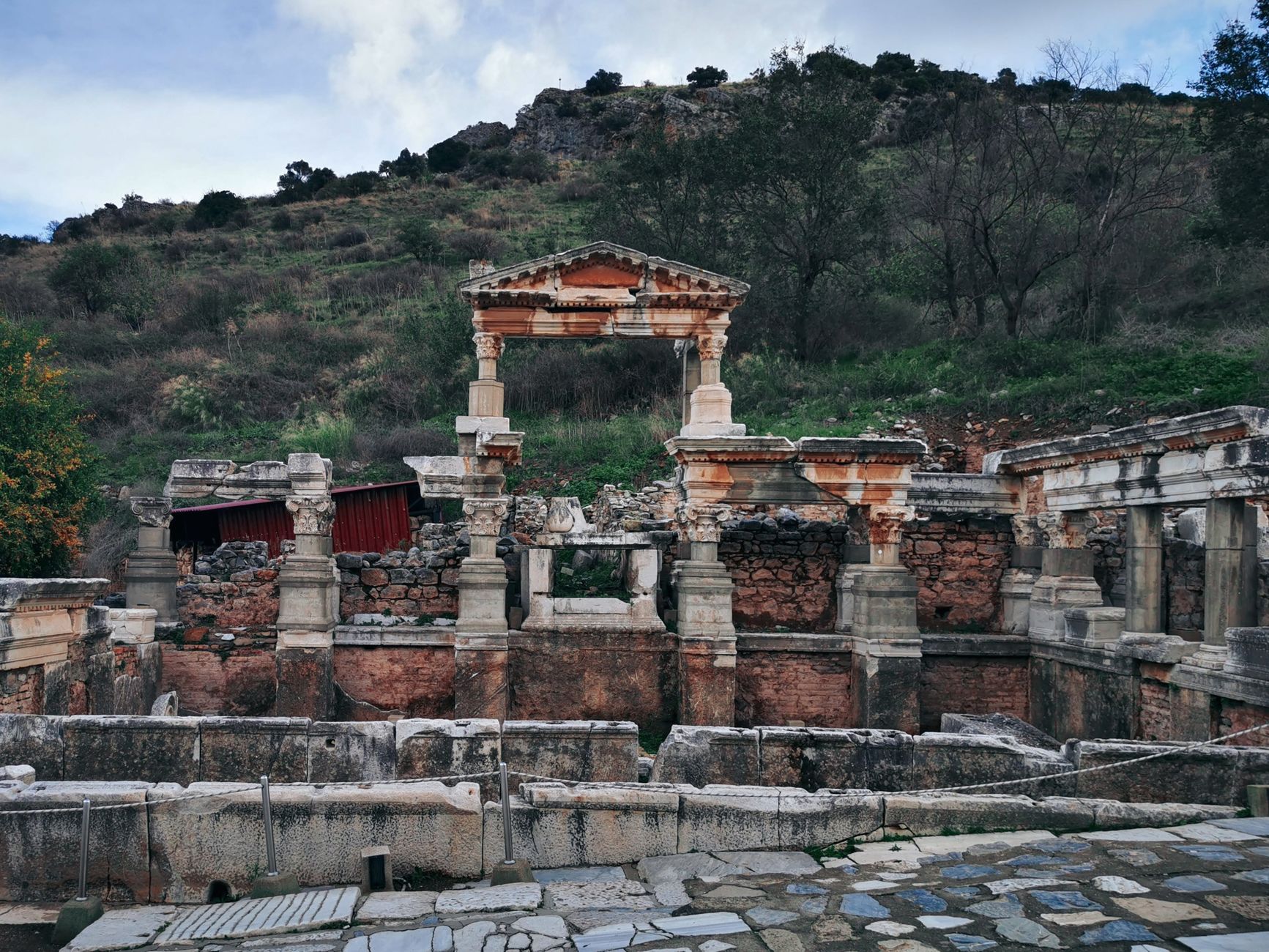 Excavated stone ruins and a standing arched fragment below a wooded hillside