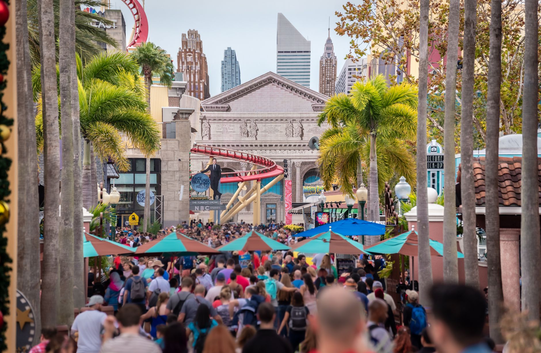 Crowds filling a palm-lined theme-park street beneath ride structures
