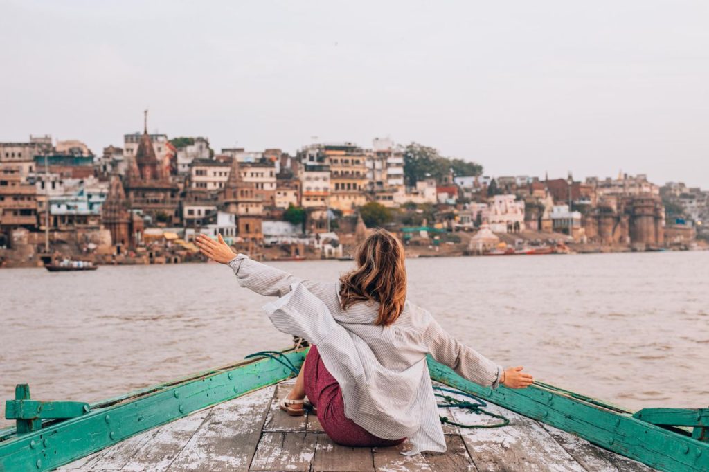 Woman sitting on a boat with arms raised, overlooking the ghats of Varanasi, India.
